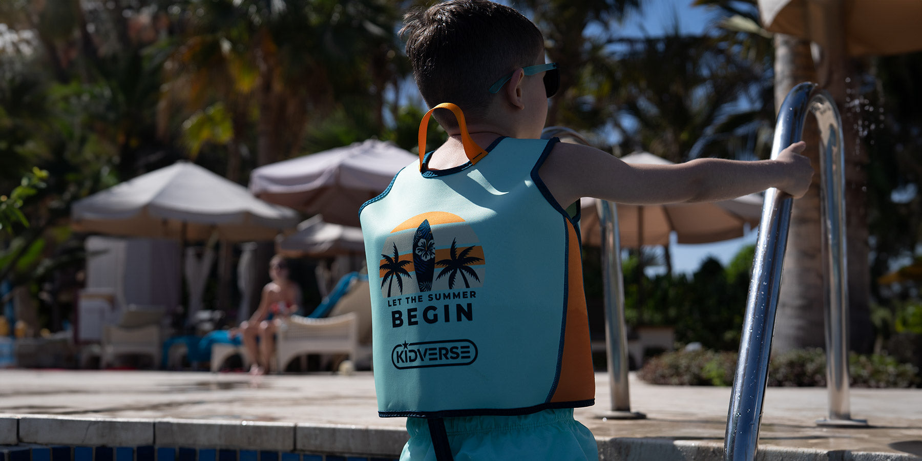 Ein Kind mit einer türkisfarbenen Schwimmweste steht am Rand eines Pools und hält sich an einer Poolleiter fest. Im Hintergrund sind Sonnenschirme, Liegen und Palmen zu sehen.