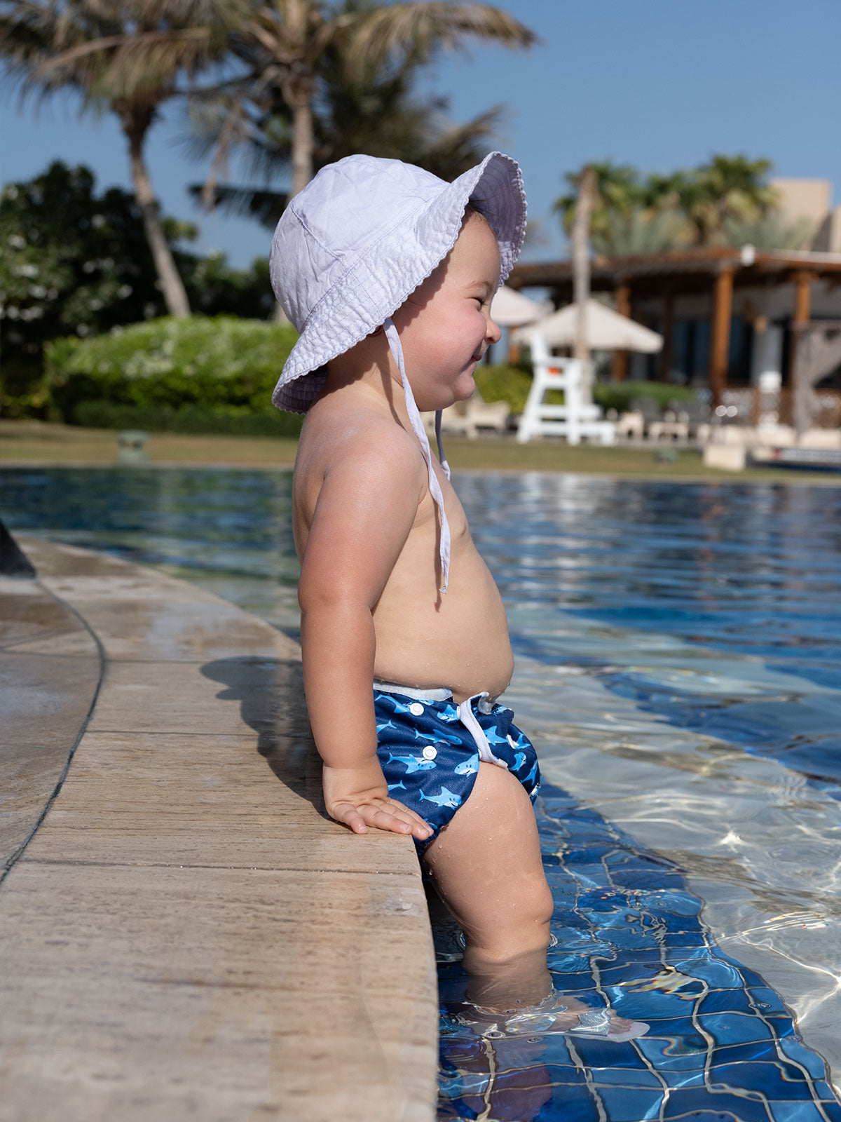 Baby sitzt am Poolrand und trägt eine blaue Schwimmwindel mit Hai-Motiven
