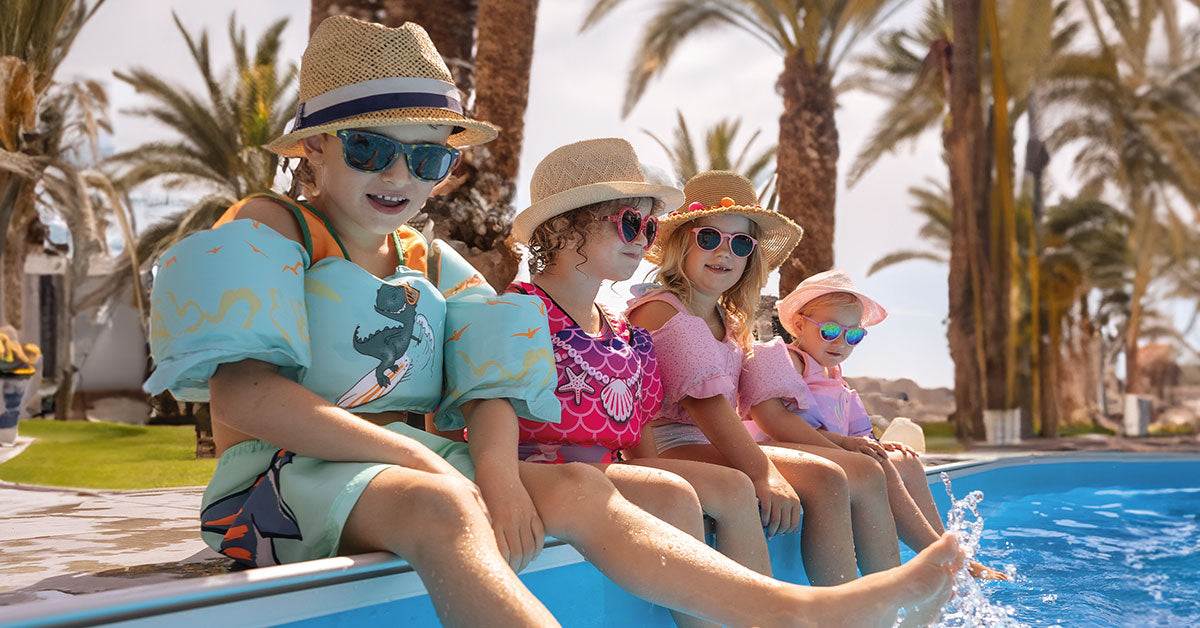 Kinder tragen Schwimmwesten mit Schwimmflügeln und sitzen am Pool, im Hintergrund sind Palmen zu sehen.