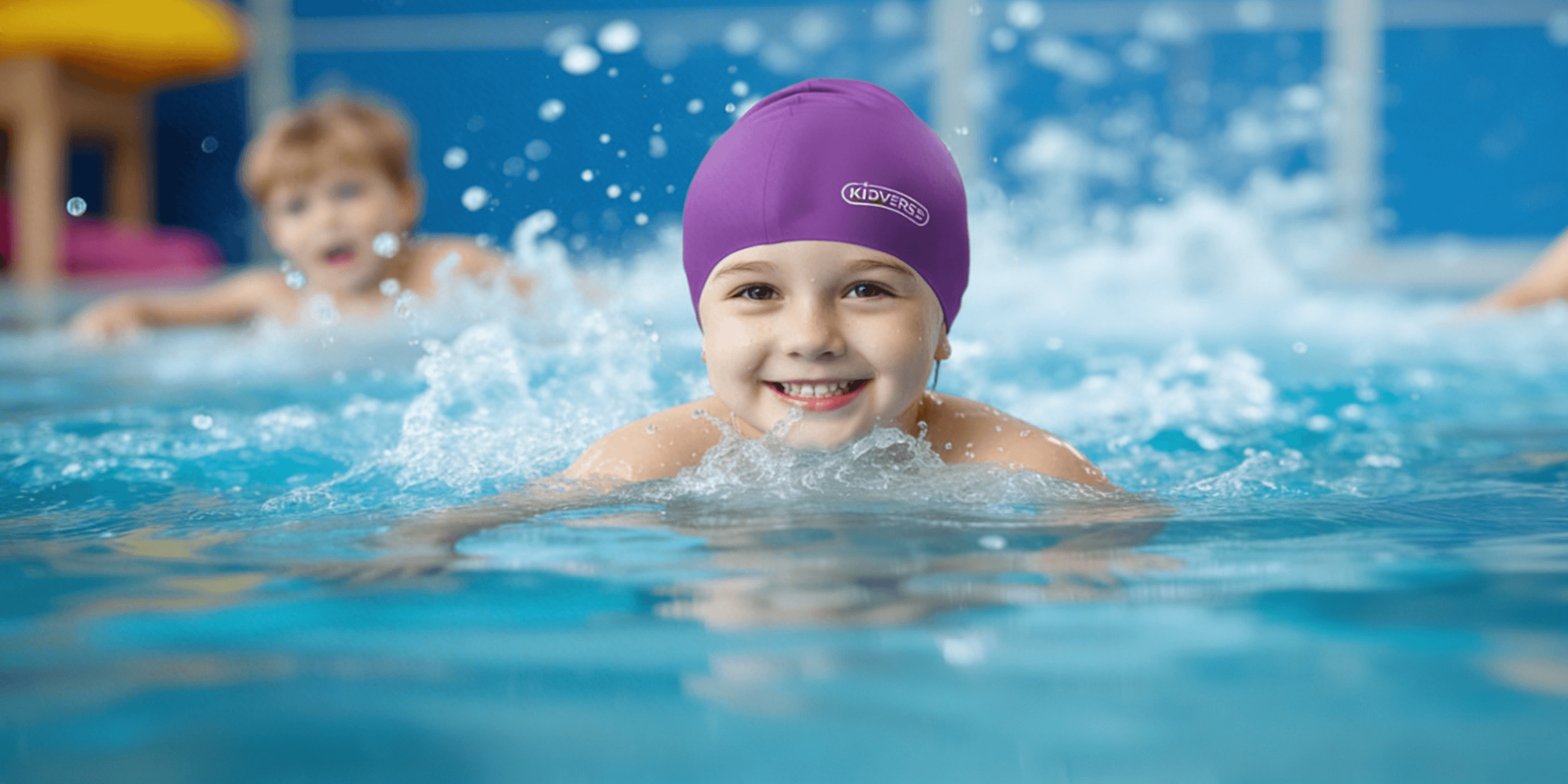 Kind im Schwimmunterricht trägt eine violette Badekappe und blickt fröhlich in die Kamera, im Hintergrund ein weiteres Kind im Wasser.